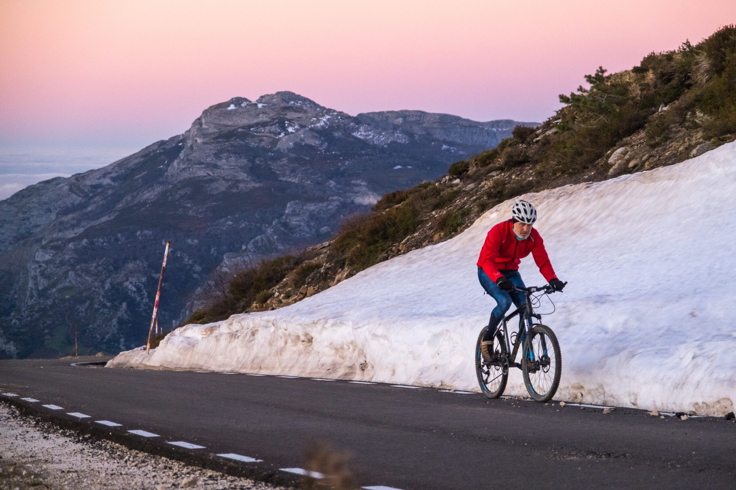 Cyclist in red jacket rides uphill on a snow-lined mountain road at sunset.